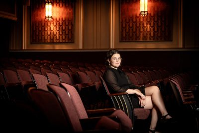 A woman sits on a seat within a row of seats in a theatre.