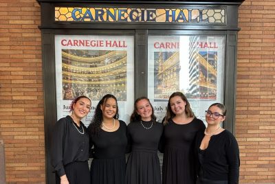 Five students in black concert attire stand together smiling outside Carnegie Hall in New York City, with the venue’s posters and stained-glass sign visible behind them.