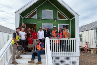 A group of community members and construction workers in safety vests stand smiling together on the porch of a newly built modular home in Maui.