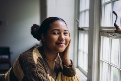 A Black young woman wearing a brown striped sweater poses near a window.